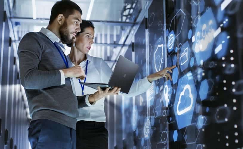 Image of a middle-aged woman and man holding a laptop in front of a touch panel displaying a visual representation of data management in cloud