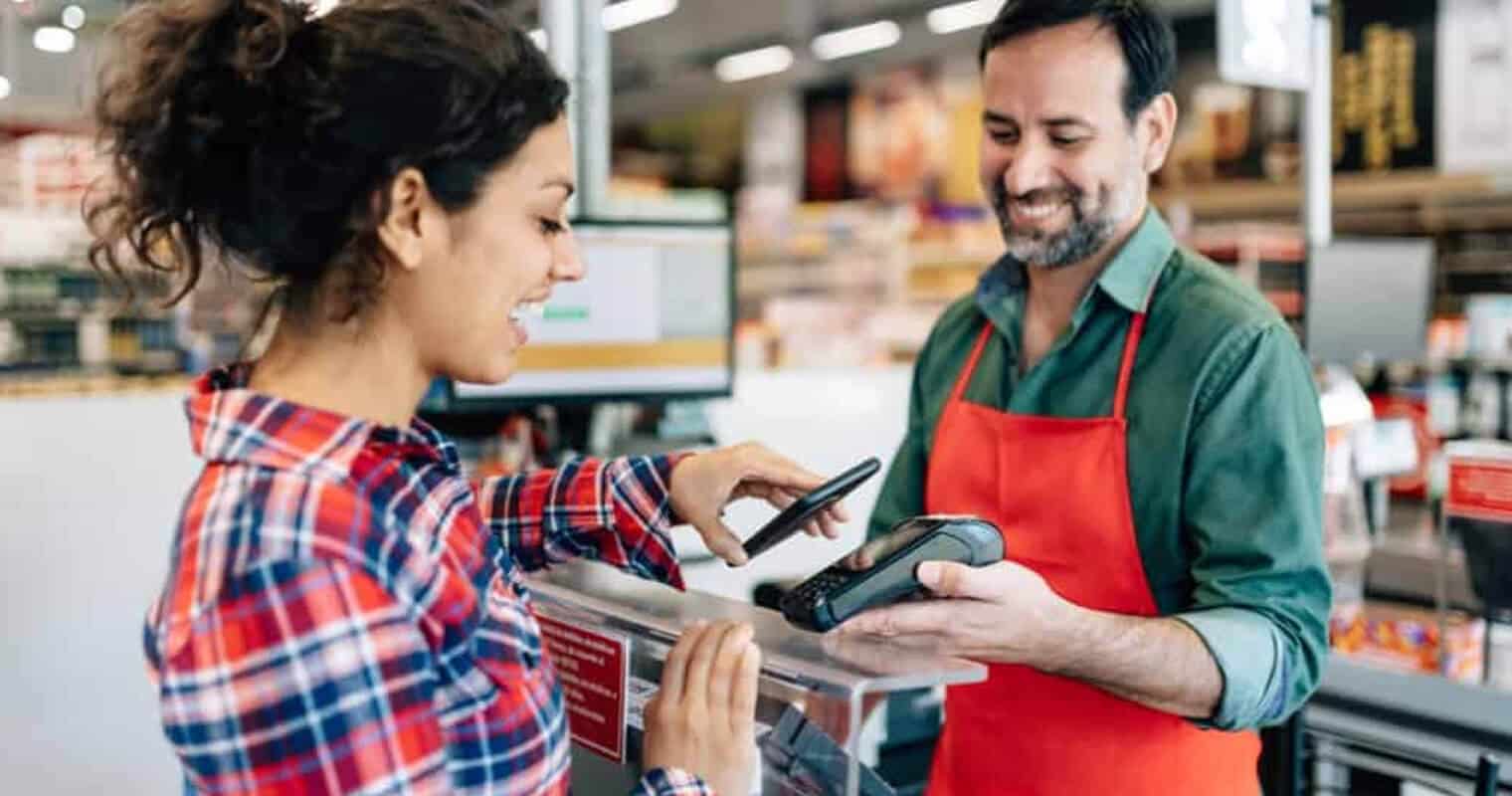 Un client paie par smartphone dans un magasin, la caissière sourit, illustrant &quot;Qu&#039;est-ce que la gestion des données par le client&quot; dans un contexte de vente au détail.