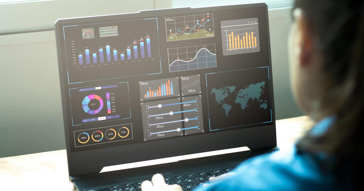 A professional is viewing a laptop screen displaying various graphs and charts that represent operational analytics data, including world maps and performance metrics.