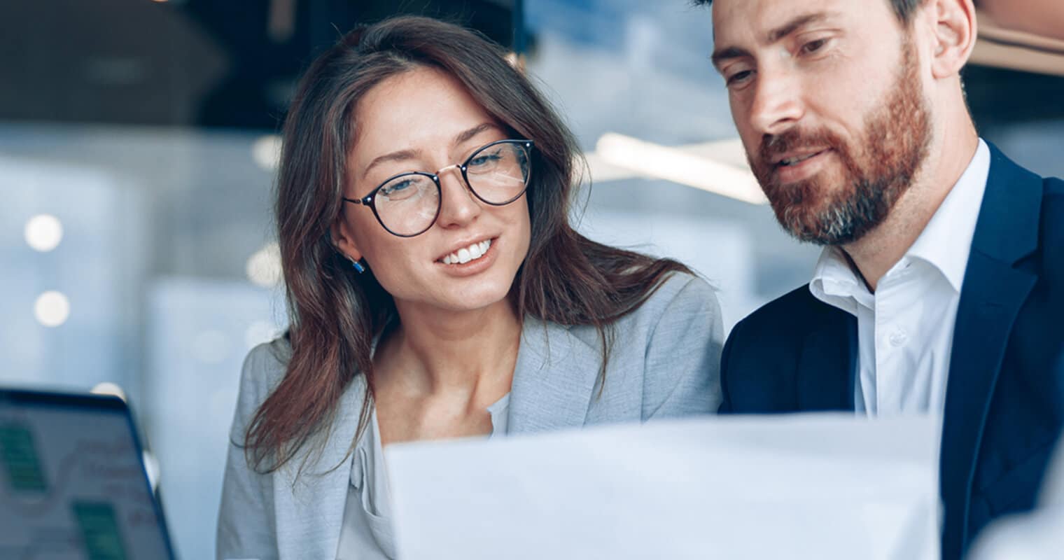 Deux professionnels, un homme et une femme, en tenue de travail, examinent ensemble un document dans un bureau moderne. La femme, qui porte des lunettes, sourit alors qu'ils discutent de la gestion des données planning.
